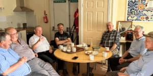7 Elderly Men sit around a table to enjoy tea and scones